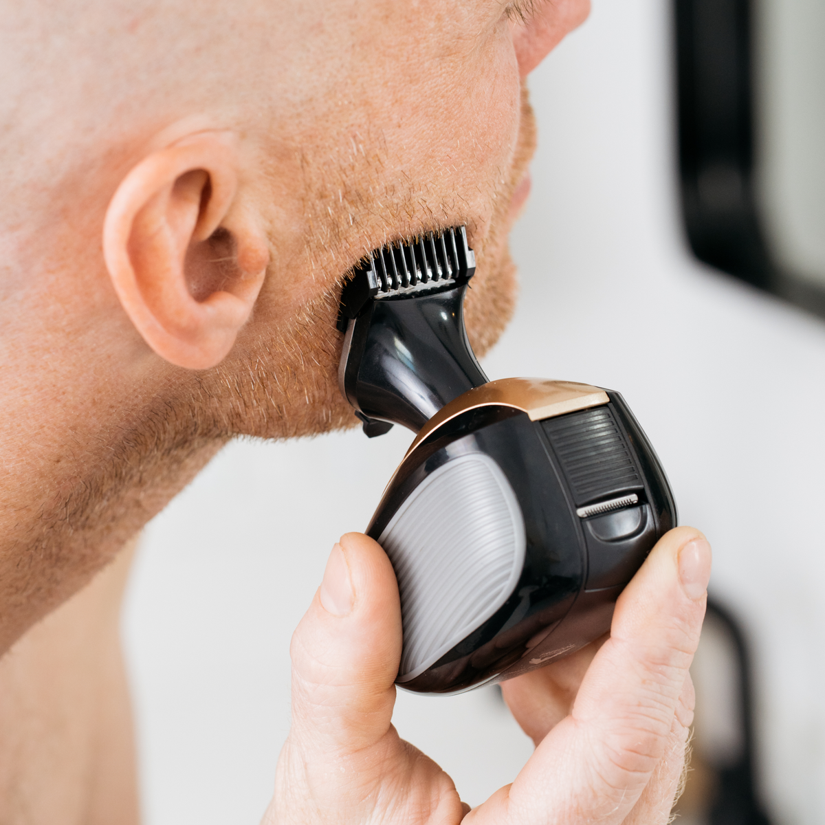 A man expertly trims his beard with a Precision Clipper & Guards, achieving a smooth finish as he stands close to the mirror.
