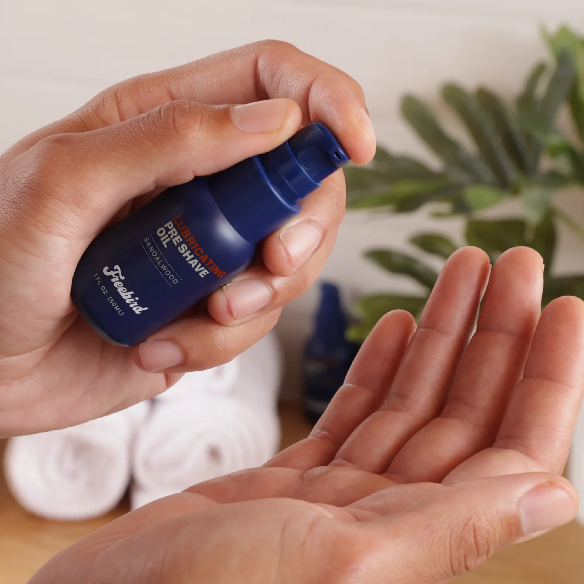 Close-up of hands dispensing Freebird's Lubricating Pre Shave Oil, infused with grapeseed oil, from a blue bottle into an open palm, with towels and a plant in the background.