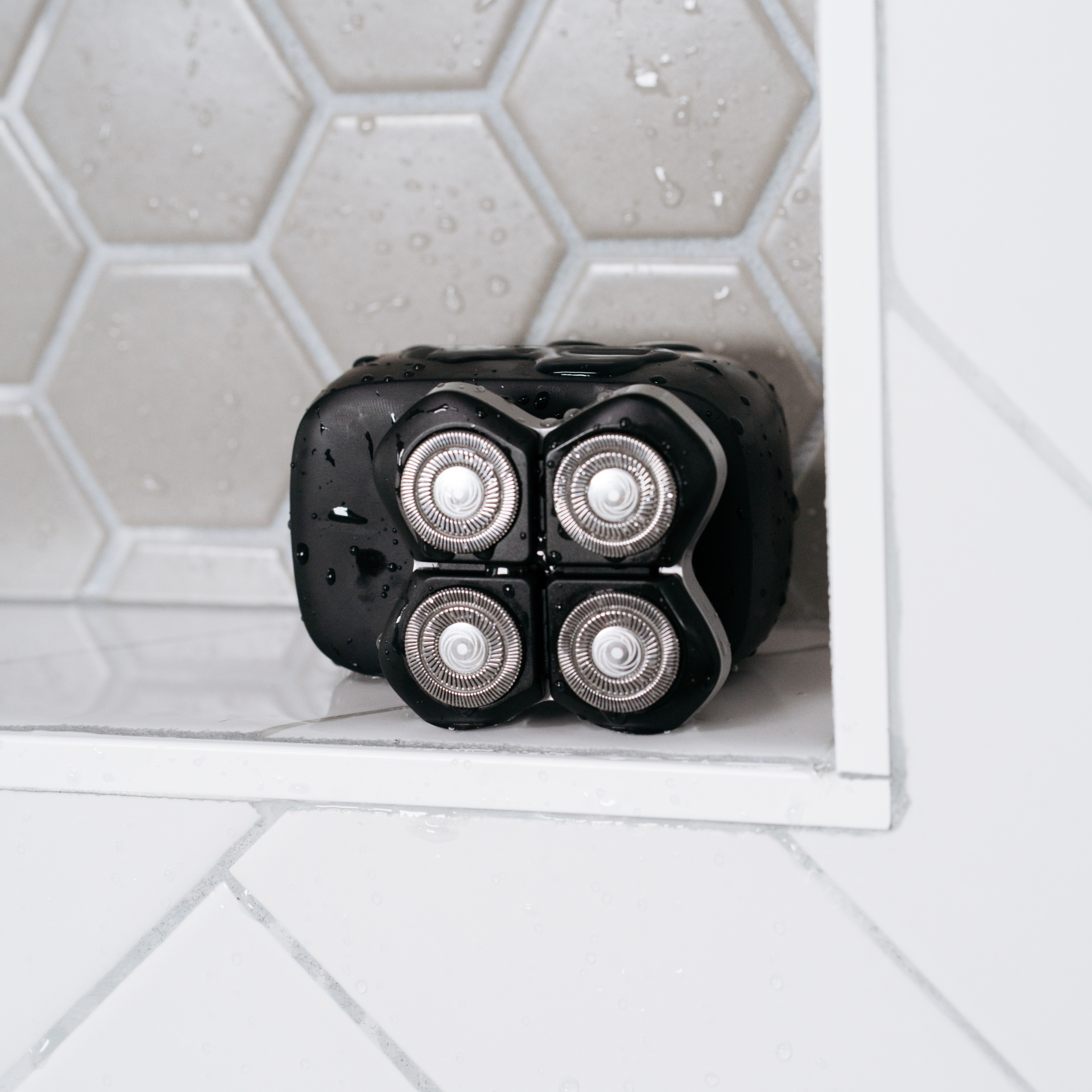 A black electric shaver with four circular blades sits on a tiled bathroom shelf with water droplets.