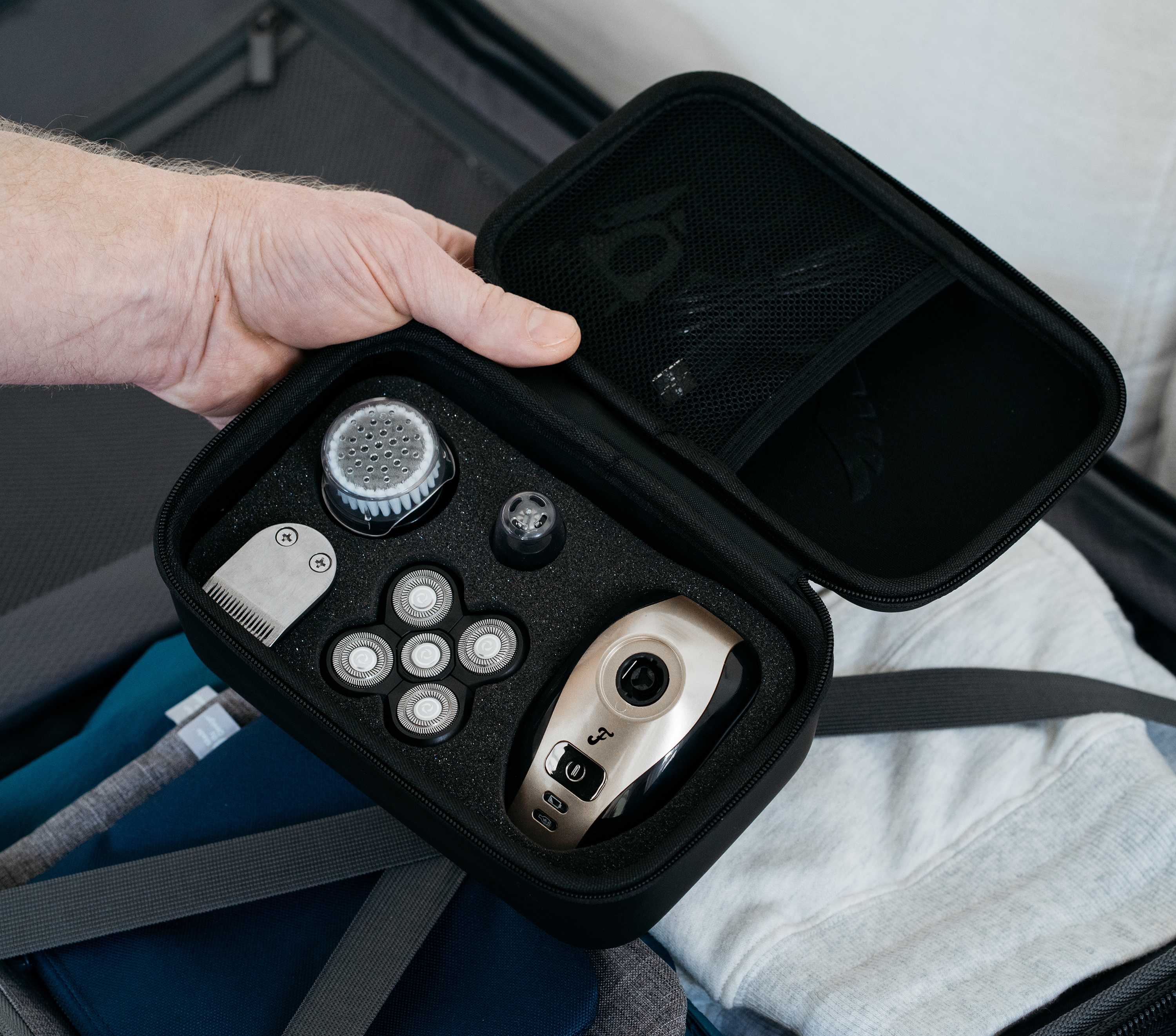 A hand holds a black Freebird FlexSeries™ Travel Case, which includes grooming tools and a free charging dock, as it rests over folded clothes in a suitcase.