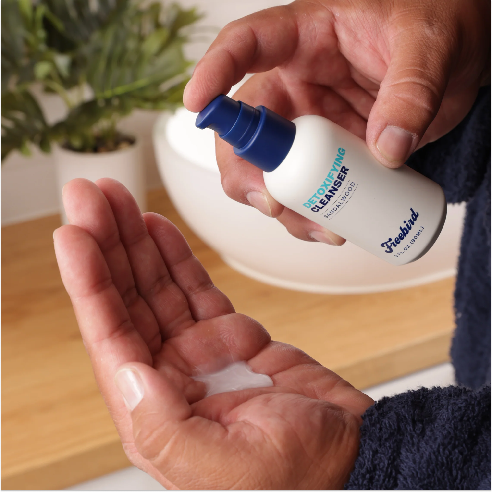 Person dispensing Freebird Detoxifying Bald Head Cleanser onto their palm, with a plant and bowl in the background, for purifying refreshment.